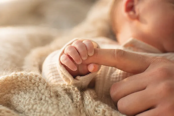 A newborn baby’s hand curled around an adult’s finger on a knitted blanket